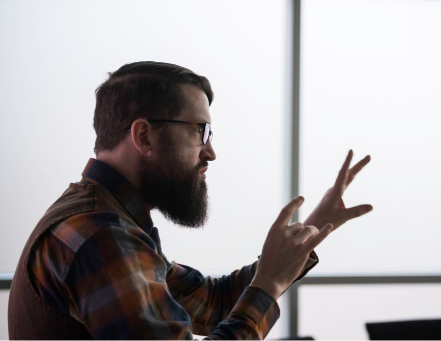 Side view profile shot of Michael Skyer. He has light skin and dark hair and beard. He is wearing classes and is communicating via American Sign Langugage